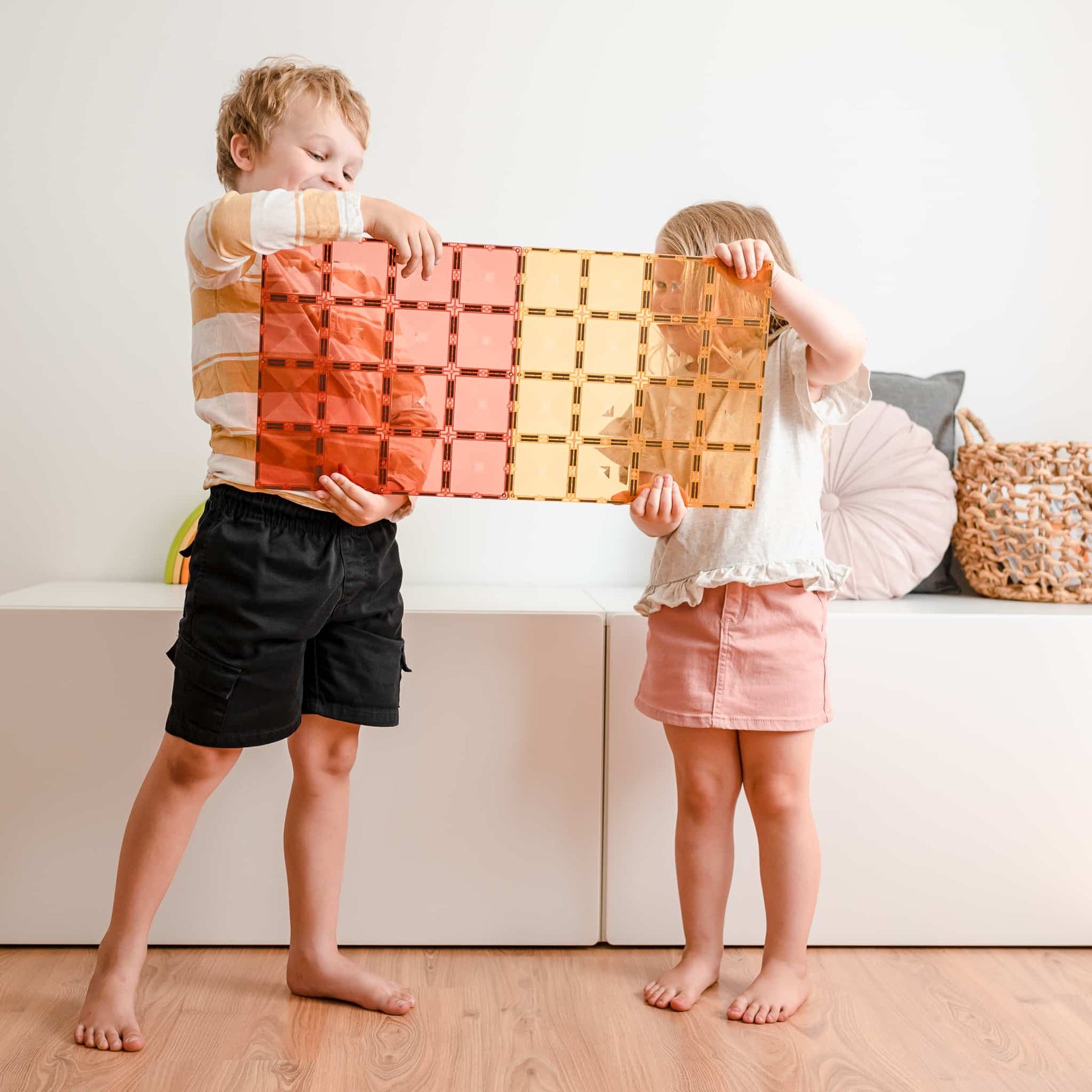 Two children holding up a colorful woven basket in a room with a white wall and wooden floor.