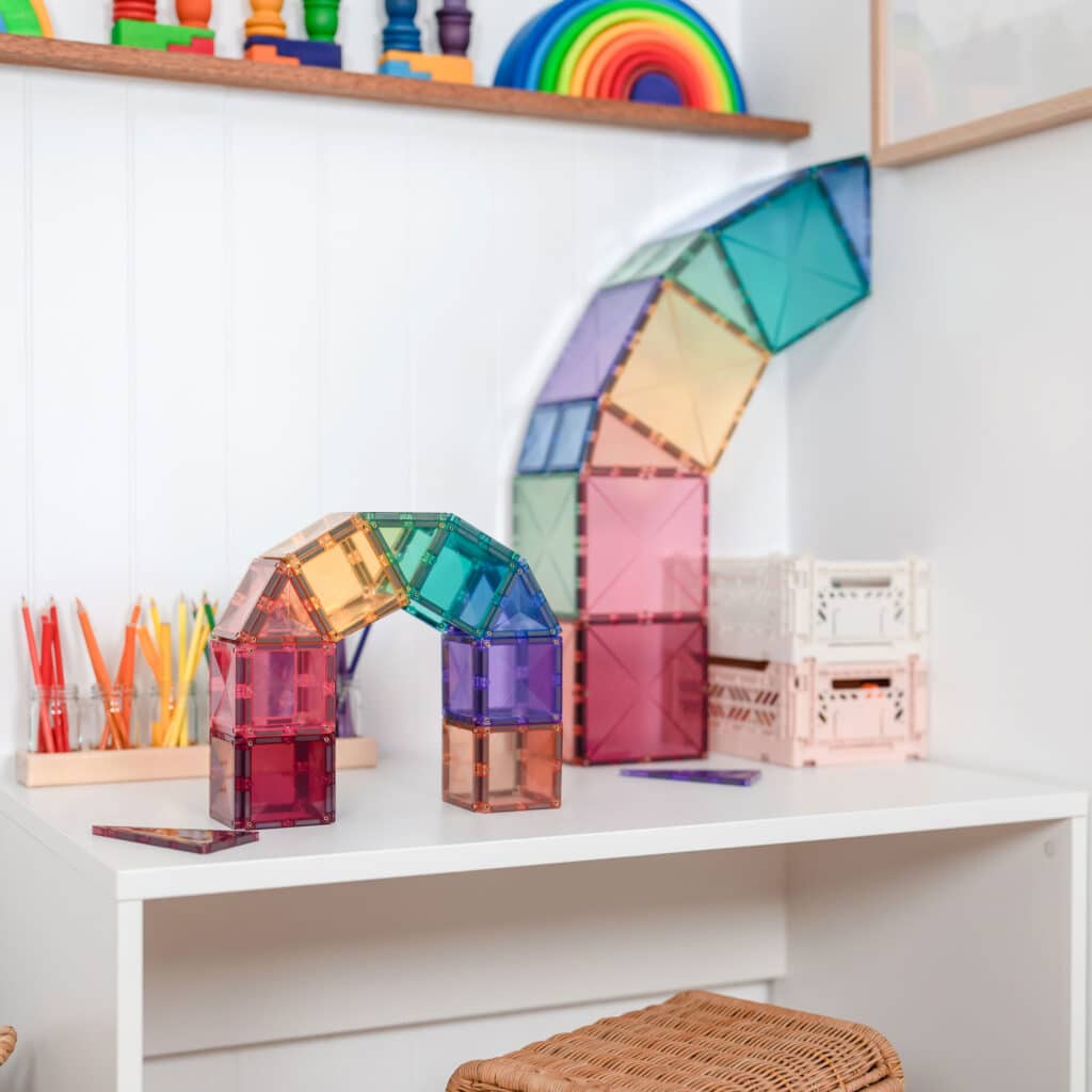 Colorful transparent magnetic building blocks on a white shelf with a rainbow-themed toy in the background.