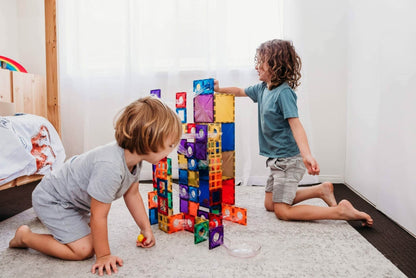 Two children playing with colorful building blocks on a carpeted floor.
