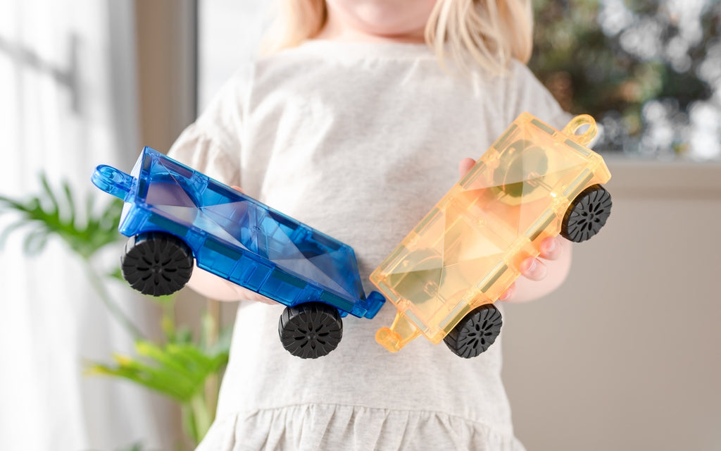 Child holding two toy cars, one blue and one orange, in front of a blurred indoor background.