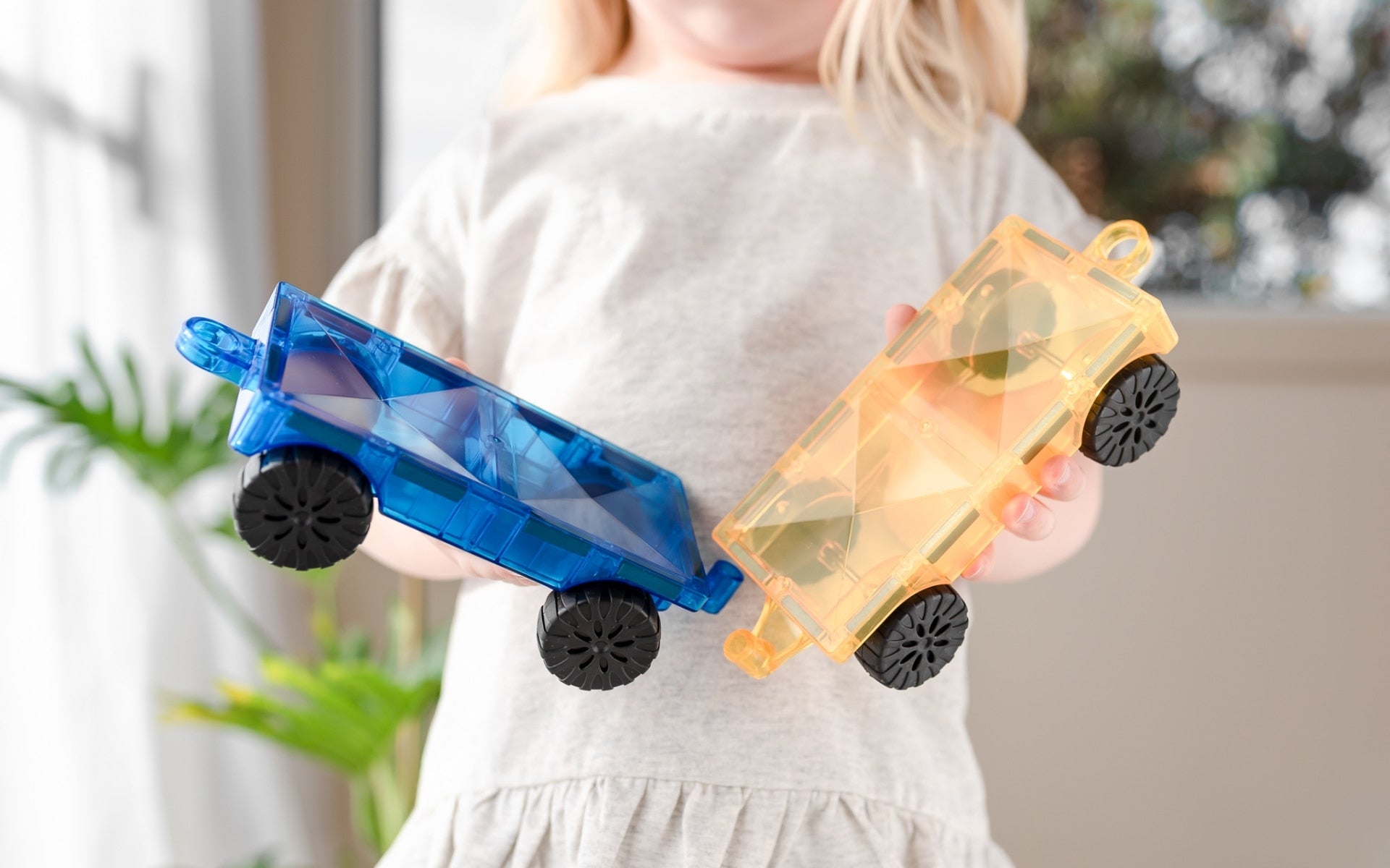 Child holding two toy cars, one blue and one orange, in front of a blurred indoor background.