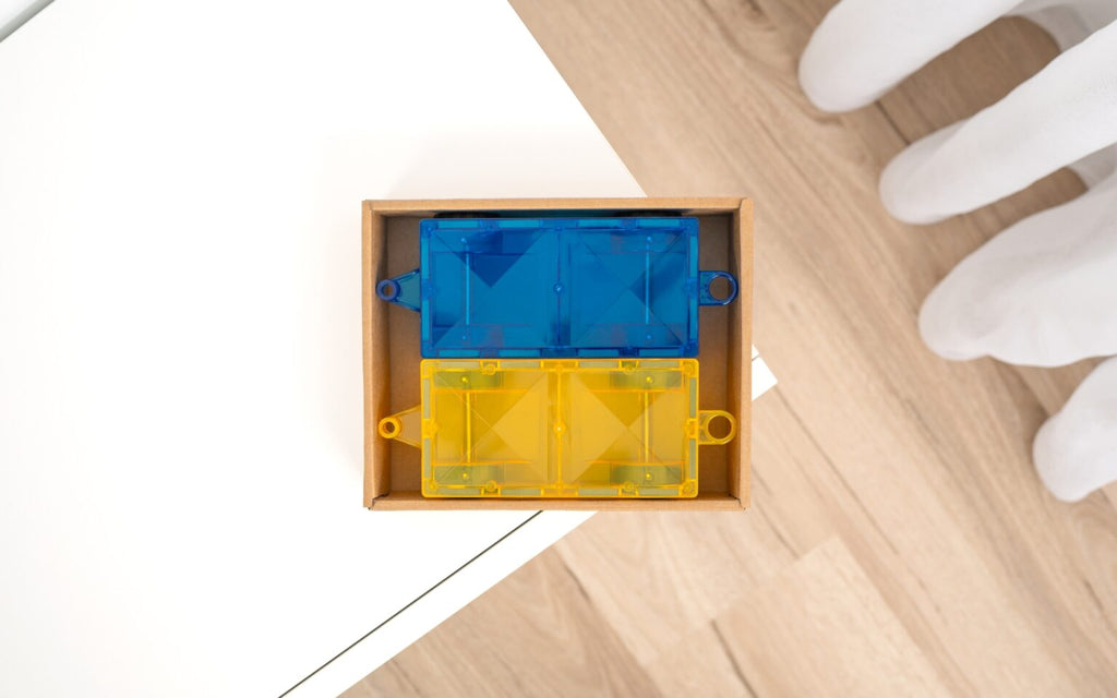Blue and yellow plastic component in a wooden box on a light wood floor with white gloves nearby.