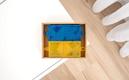 Blue and yellow plastic component in a wooden box on a light wood floor with white gloves nearby.