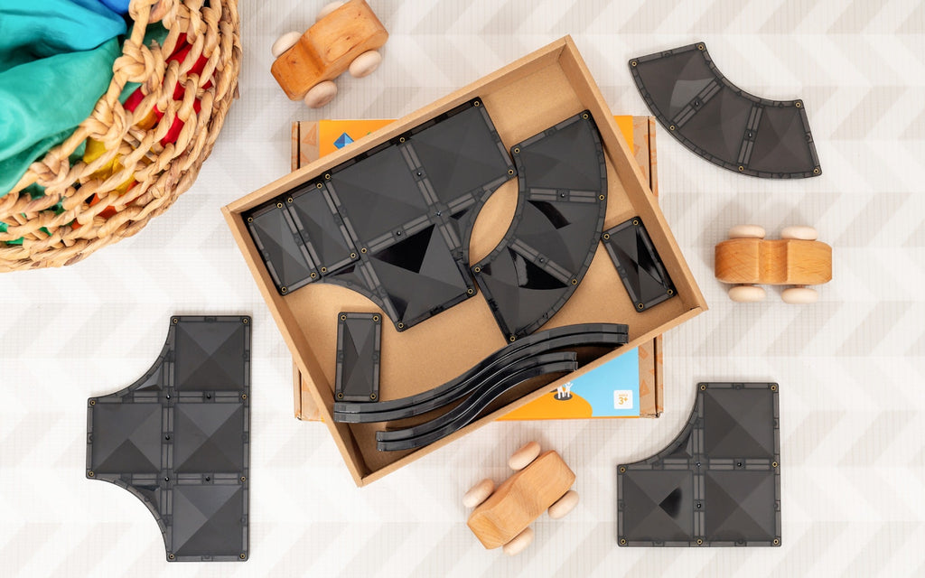 Black geometric toys in a cardboard box on a light surface with wooden toys around.