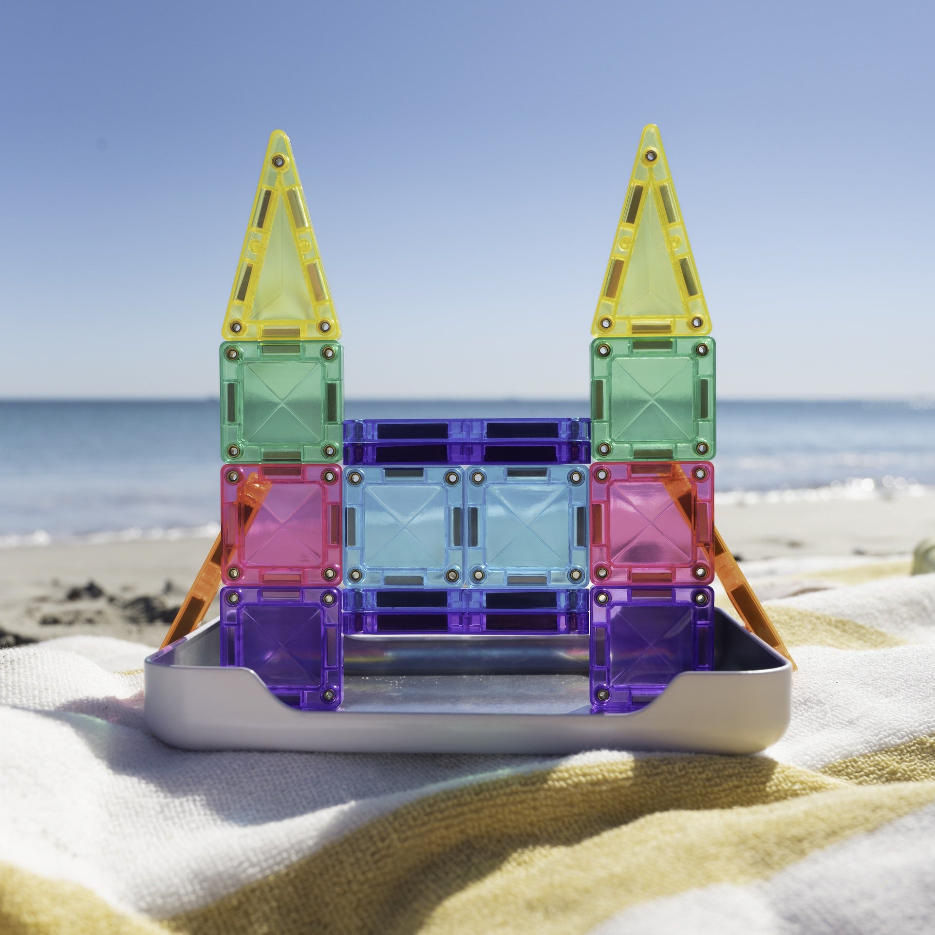 A colorful magnetic tile castle toy built in a silver tray, placed on a beach towel with the ocean and blue sky blurred in the background.