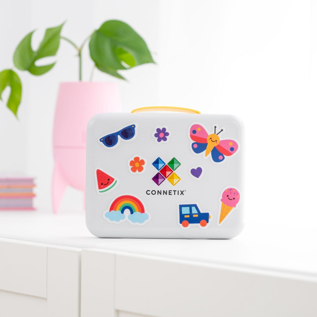 A white Connetix box with a yellow handle, decorated with colorful cartoon stickers including a butterfly, rainbow, sunglasses, and watermelon, sitting on a white shelf with a pink planter in the background.
