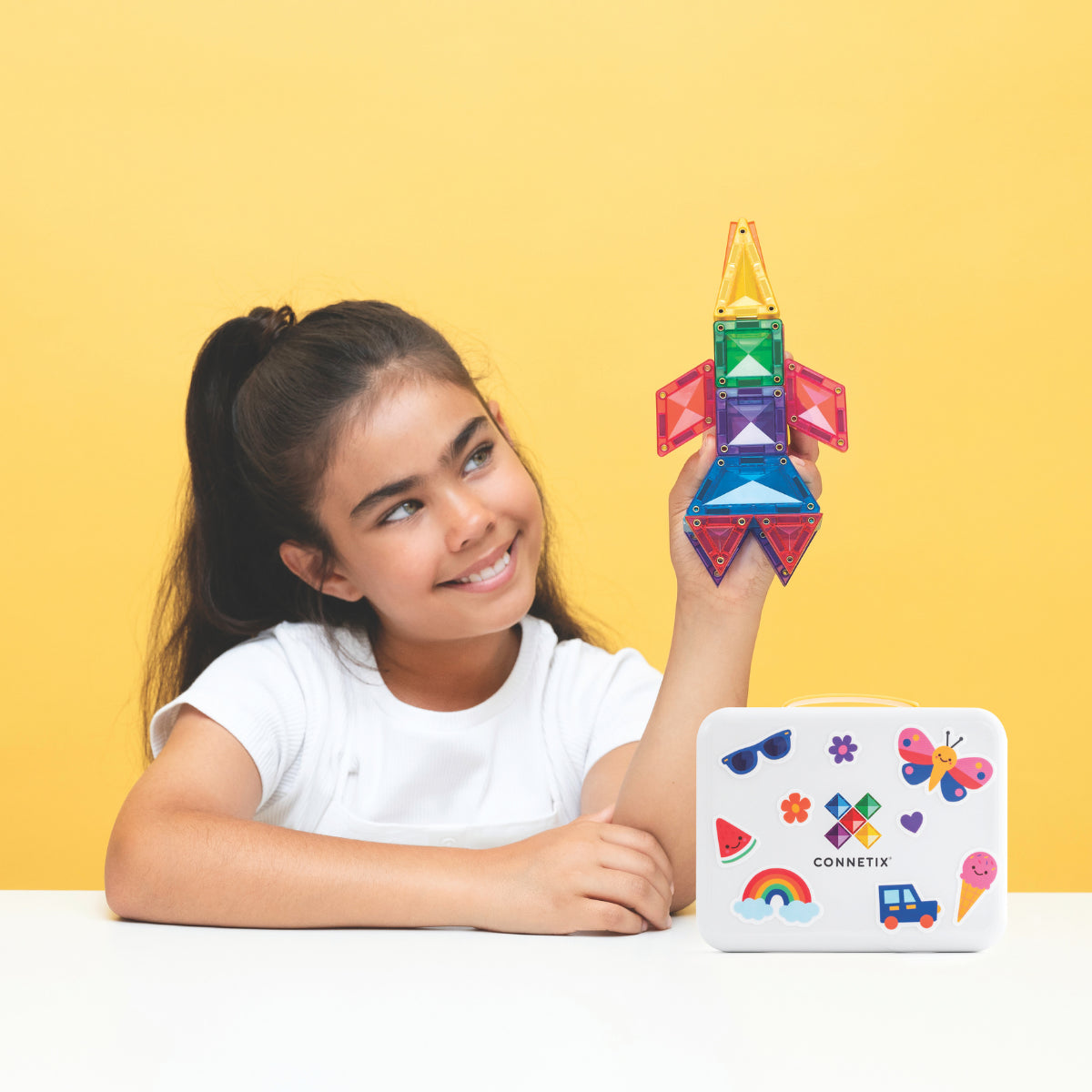 A young girl with her hair in a ponytail smiles while holding a colorful magnetic building rocket. She is sitting at a white table against a solid yellow background, with a white Connetrix storage box featuring colorful stickers placed in front of her.