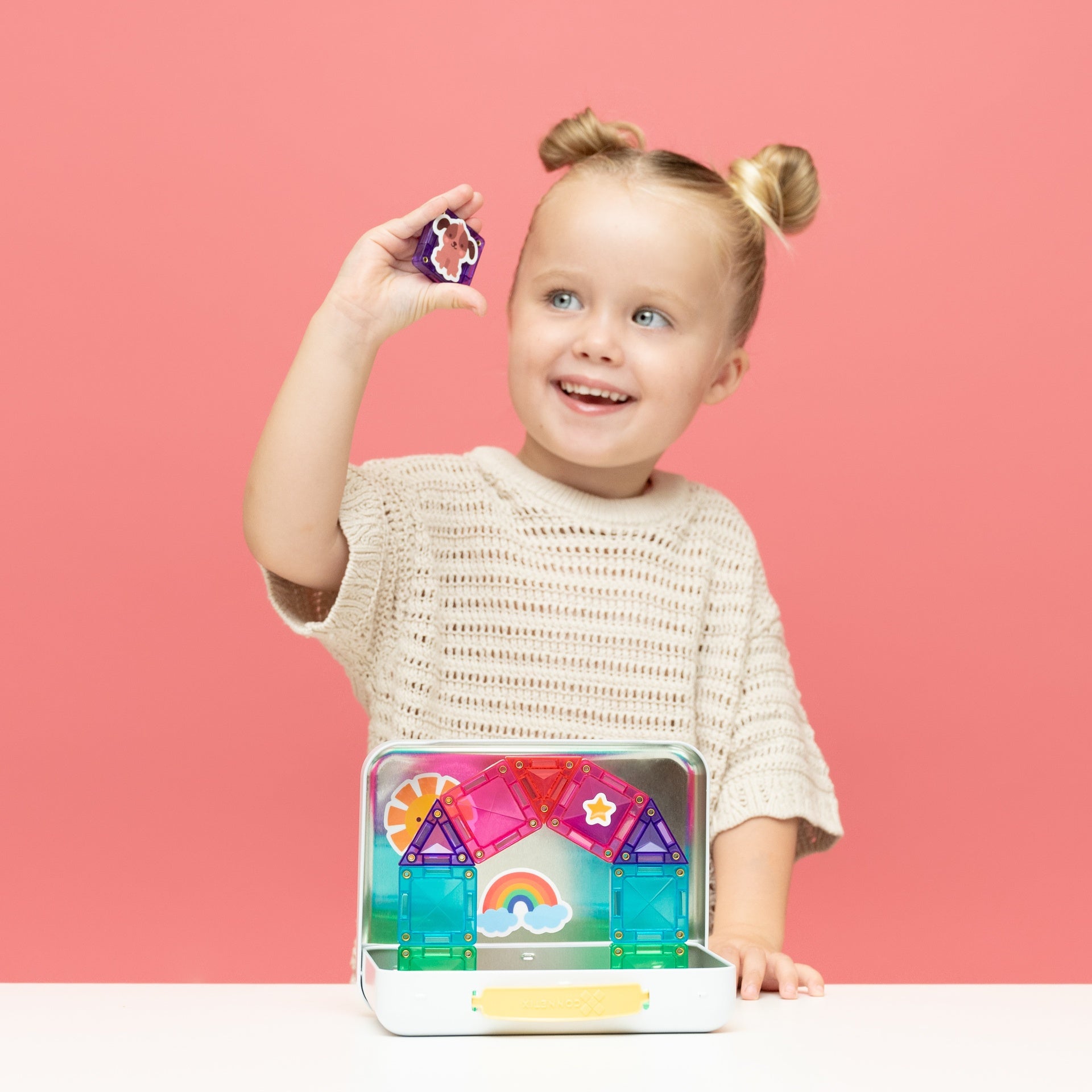A young child with blonde pigtails stands behind a table against a pink background, holding a small colorful magnetic building block above an open white case containing translucent geometric toy pieces.