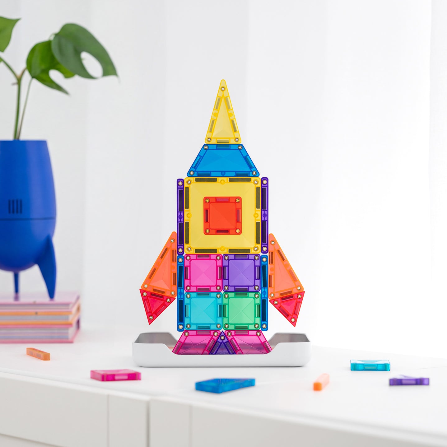 A colorful magnetic tile rocket toy built on a white display base, set on a white tabletop with a blue plant pot and green leaves in the background.