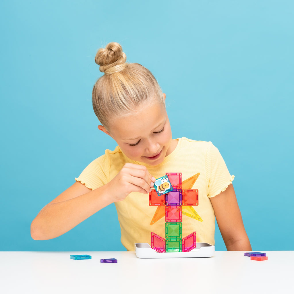A young blonde girl with her hair in a bun is sitting at a white table, building a colorful magnetic block tower with a small blue and yellow character piece. She is wearing a yellow t-shirt against a solid light blue background.