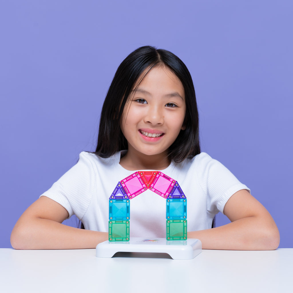 A young girl with black hair smiles while sitting at a white table, wearing a white t-shirt and posing with a colorful magnetic arch toy built from translucent tiles on a white base against a solid purple background.