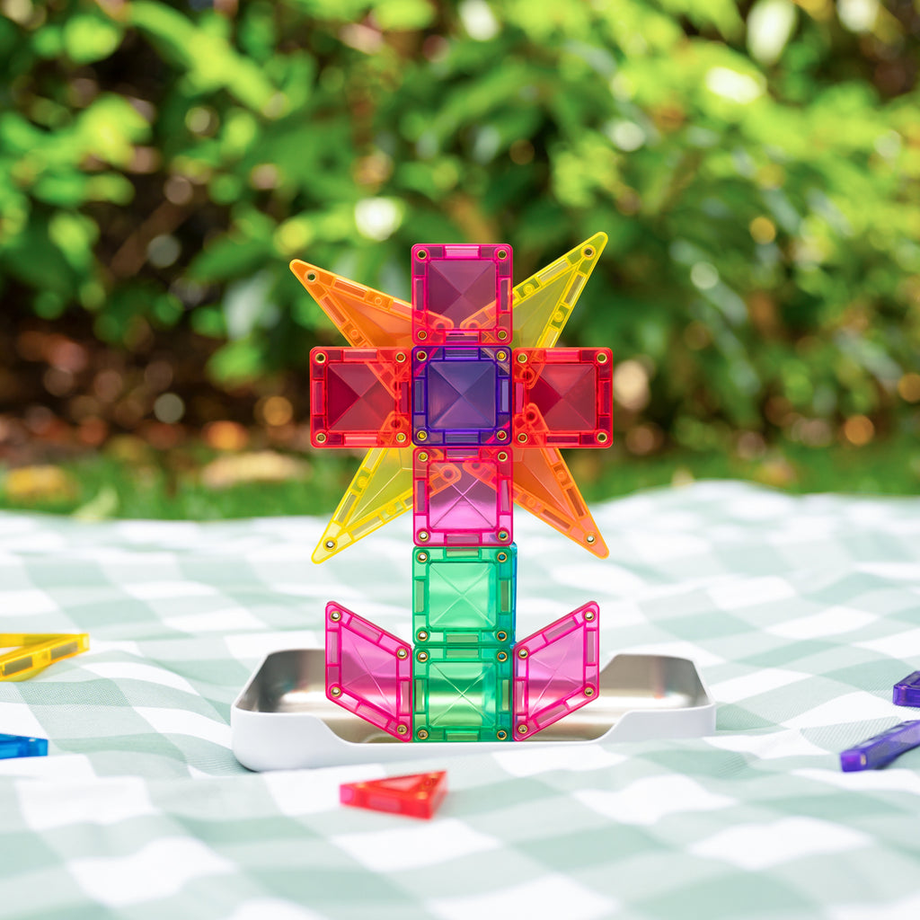 A colorful translucent magnetic building block toy structure stands on a small metal base on a white checkered blanket outdoors, with blurred green foliage in the background.