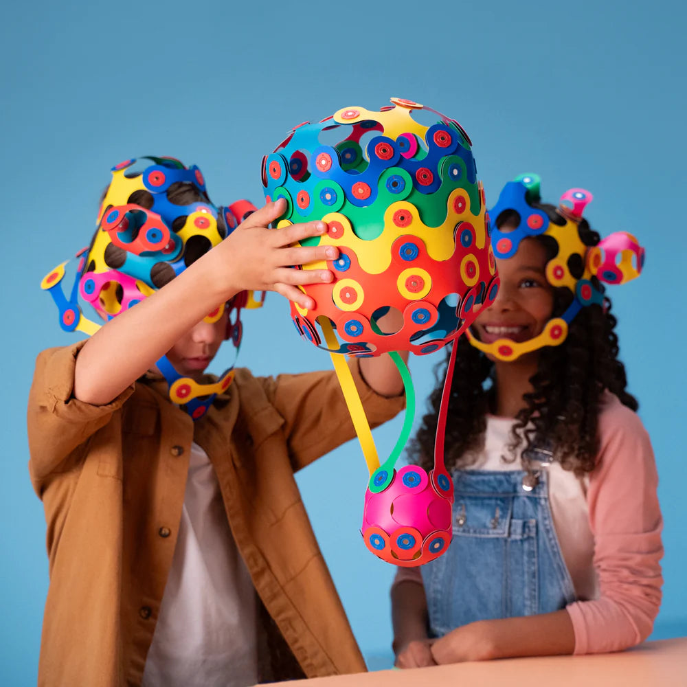 Two children wearing colorful helmets with a large multicolored helmet in the center against a blue background.
