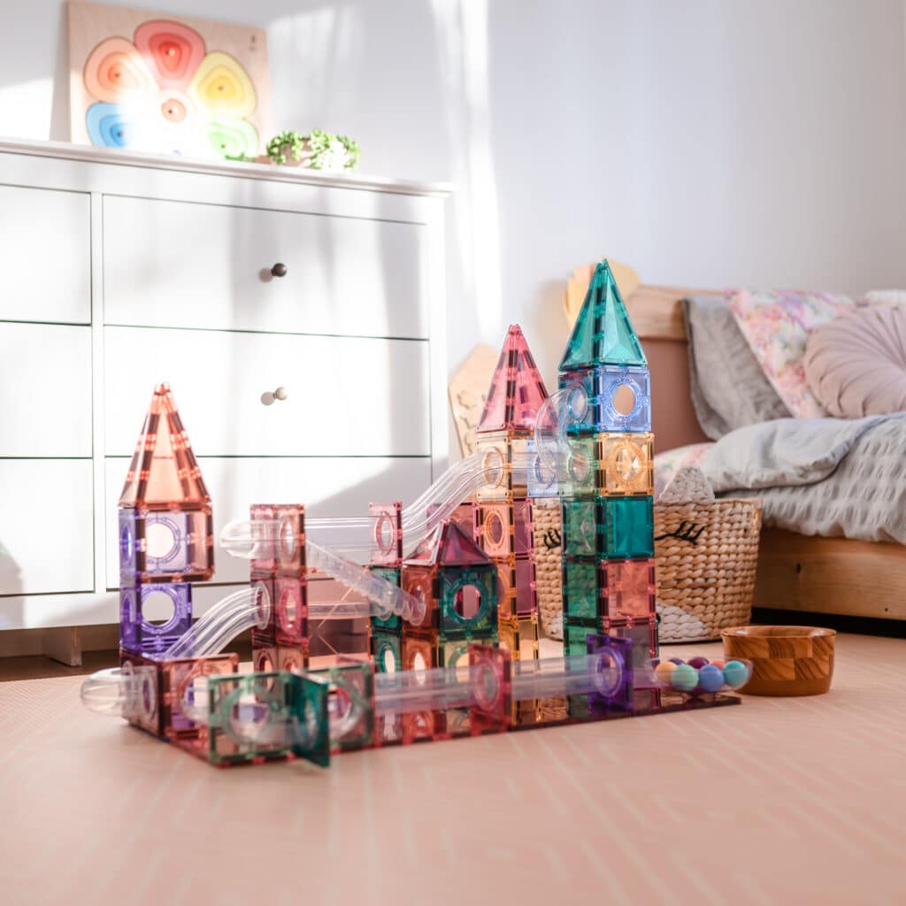 Colorful magnetic building blocks on a wooden floor with a bed and dresser in the background.