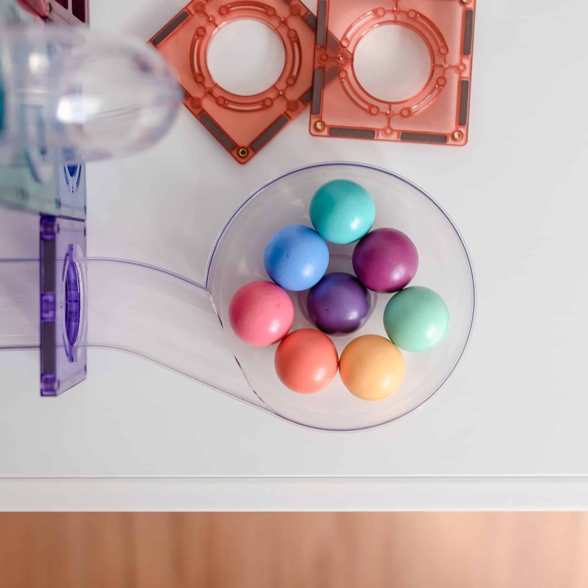 Colorful balls in a transparent container on a white surface with pink geometric shapes in the background.
