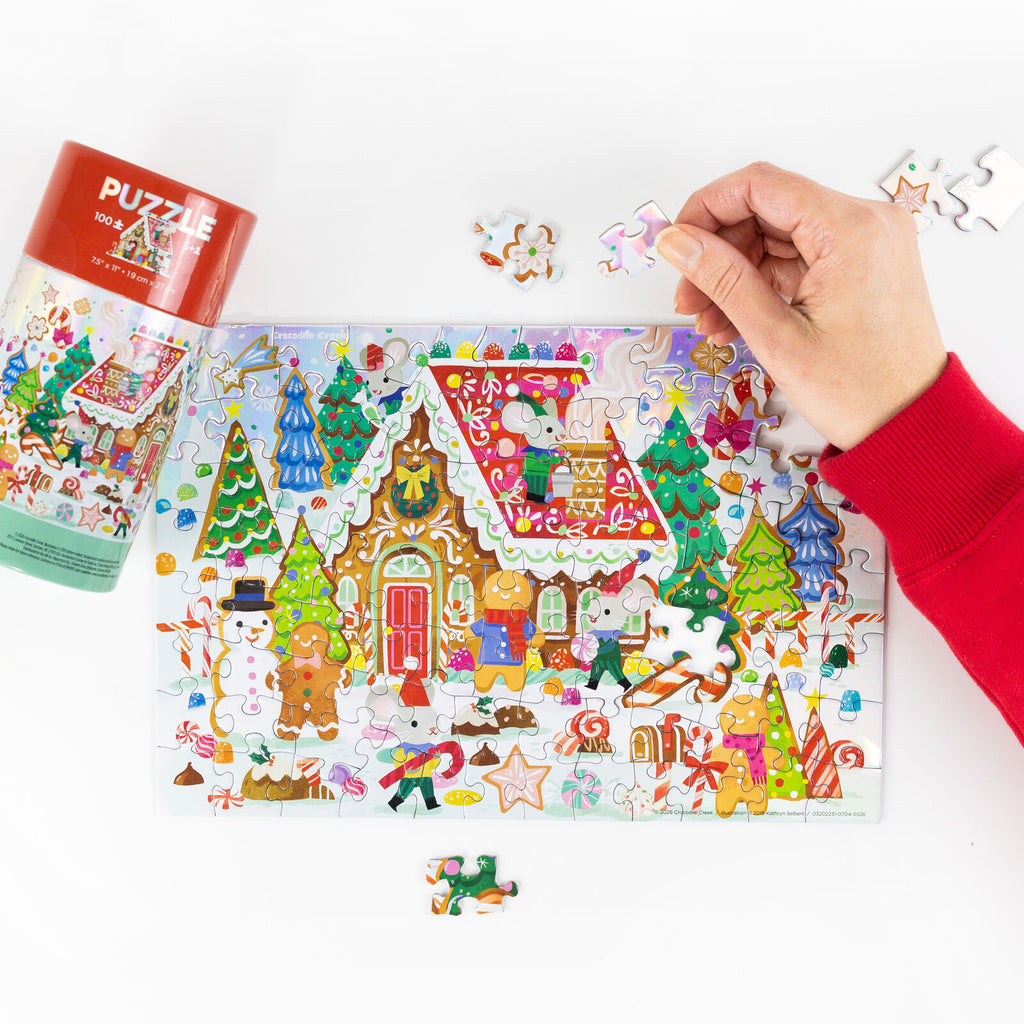 A colorful Christmas-themed jigsaw puzzle with a red cylindrical container and loose pieces on a white surface, with a person's hand placing a piece.