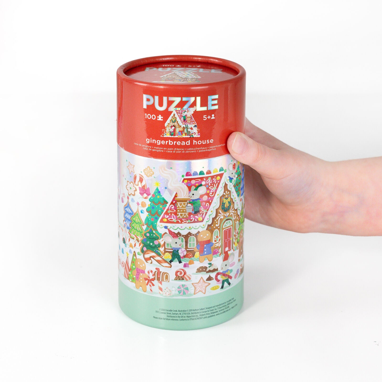 A hand holding a cylindrical puzzle box for a gingerbread house jigsaw puzzle. The box has a red top section with white 'PUZZLE' text and a colorful illustrated lower section showing holiday gingerbread scenes, on a clean white background.