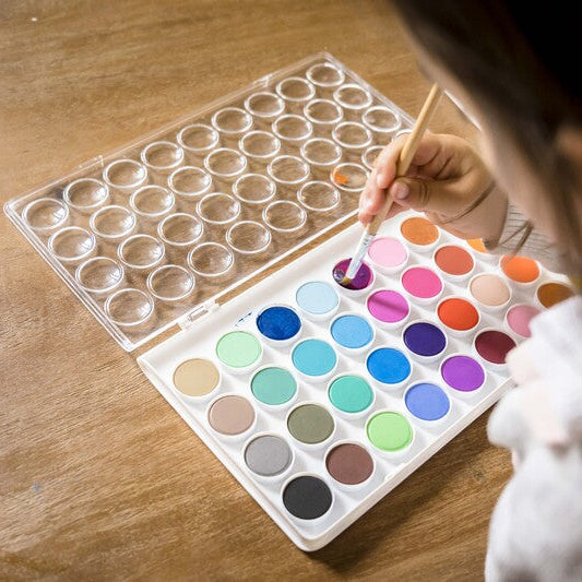 Person using a paintbrush to select a color from a watercolor palette on a wooden surface