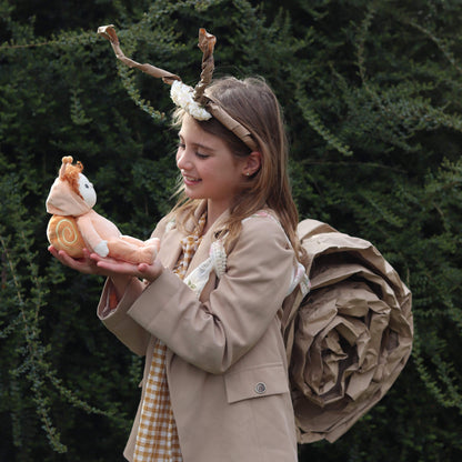 Person in a costume with antlers and a snail plush toy, standing against a green bush background.