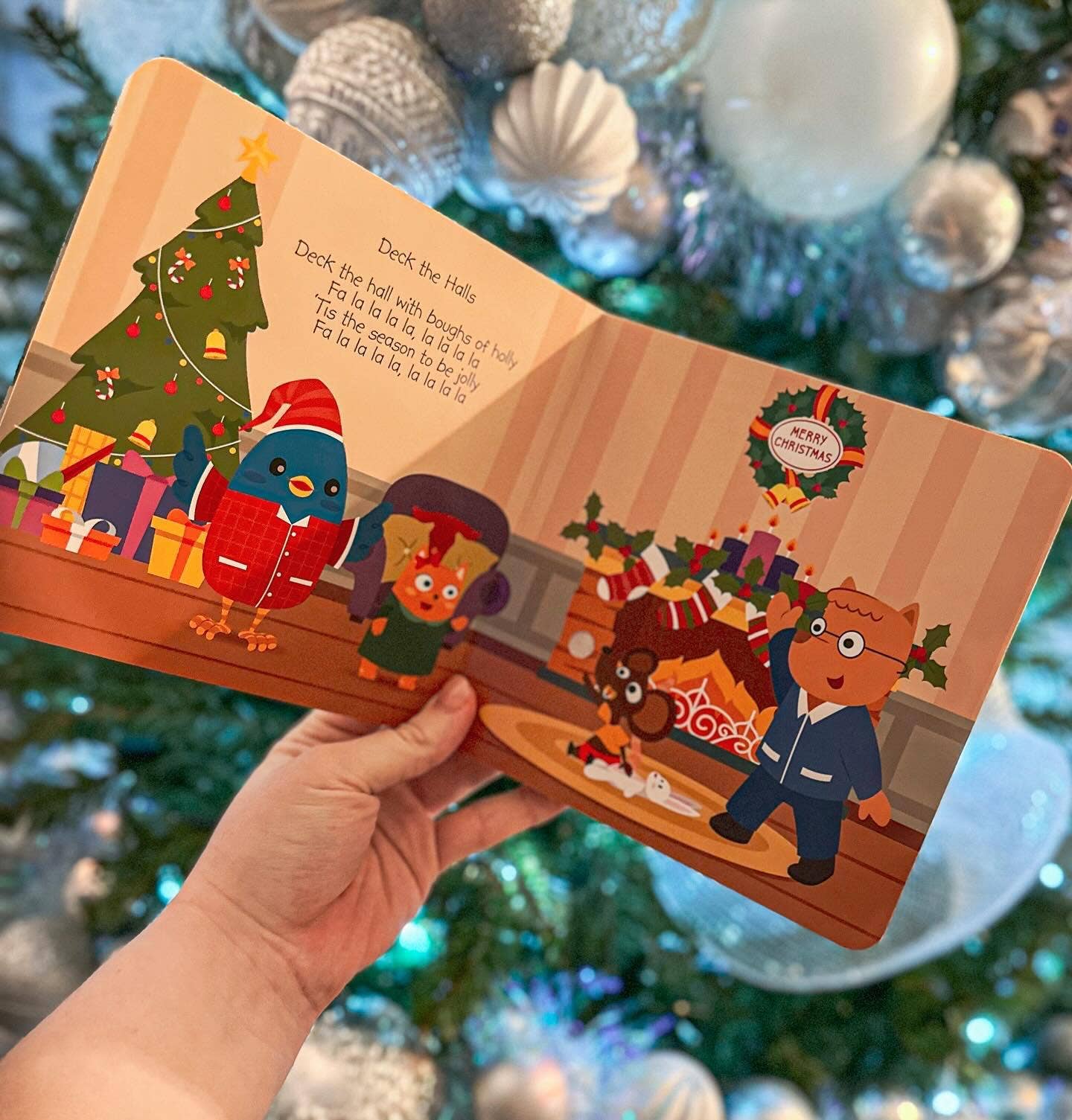 Children's book with Christmas-themed illustrations held in front of a decorated Christmas tree.