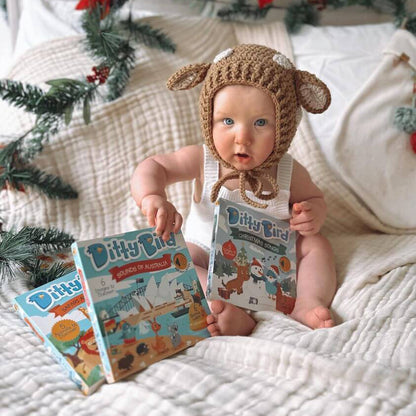 Baby in a bear hat holding books on a bed with Christmas decorations.