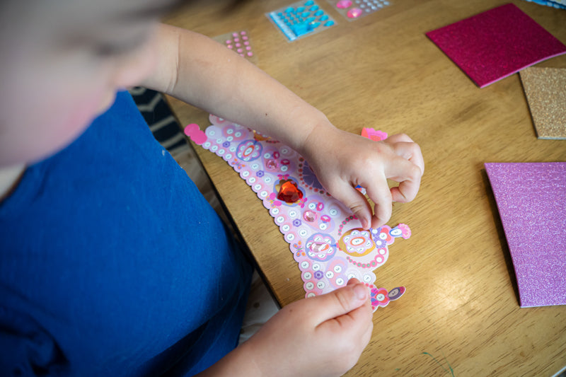 Child putting stickers on a paper tiara at a table. 
