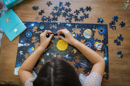 Person assembling a puzzle with space-themed pieces on a wooden table.