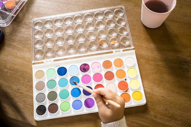 Colorful watercolor palette on a wooden table with a hand holding a paintbrush.