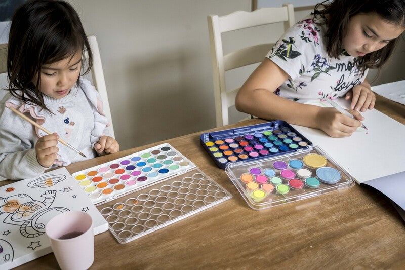 Two children sitting at a table with art supplies, including watercolor paints and brushes.