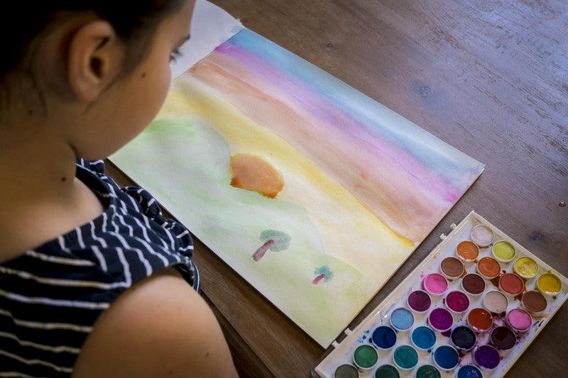 Person painting a watercolor scene of a beach with a palette on a wooden table