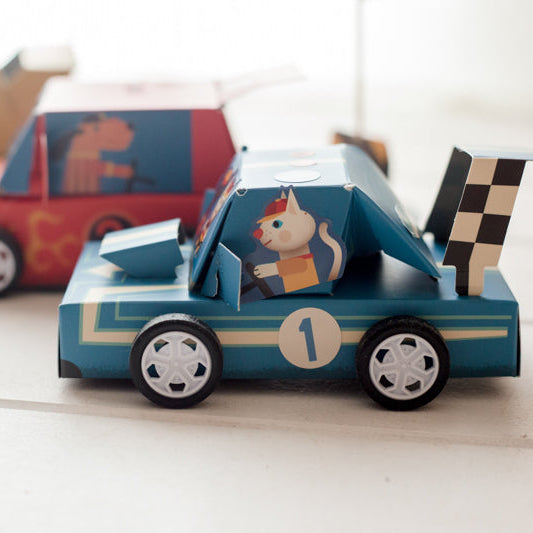 Cardboard toy car with a cat figure inside, surrounded by other toy cars on a light surface.