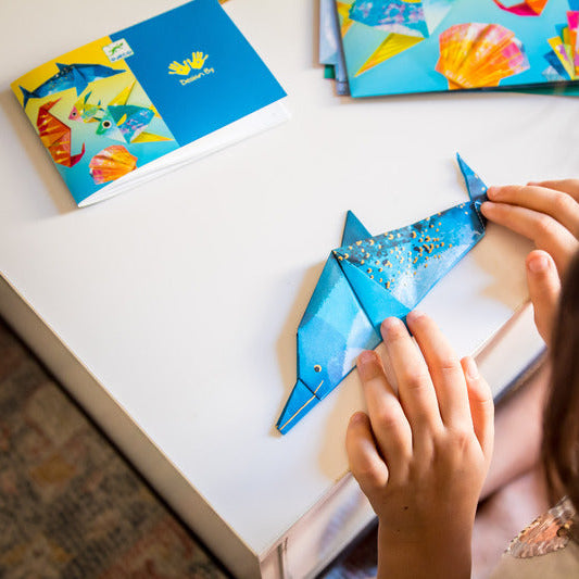 Person holding a blue origami dolphin with colorful cards in the background