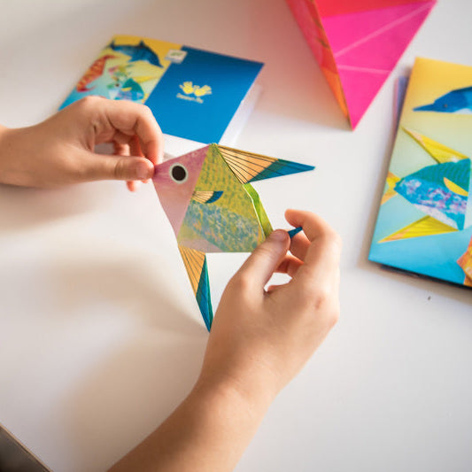 Child's hands holding a colorful paper airplane with more paper airplanes on a light surface.