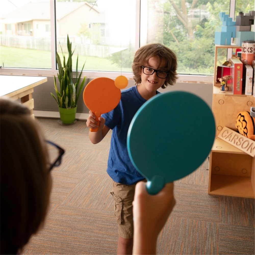 Two children playing with colorful paddles in a room with large windows and furniture.