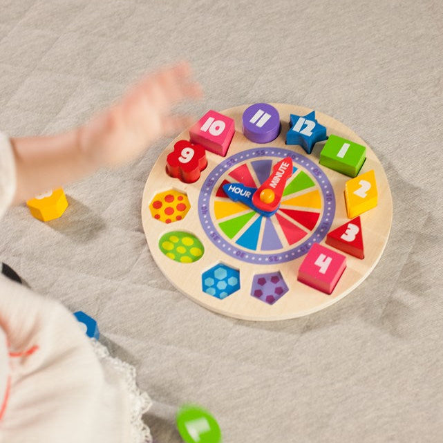 A baby's hand reaches toward a colorful wooden shape-sorting and number learning toy on a light gray fabric floor. The toy features a circular board with numbered geometric shapes in red, yellow, green, blue, purple, and orange.