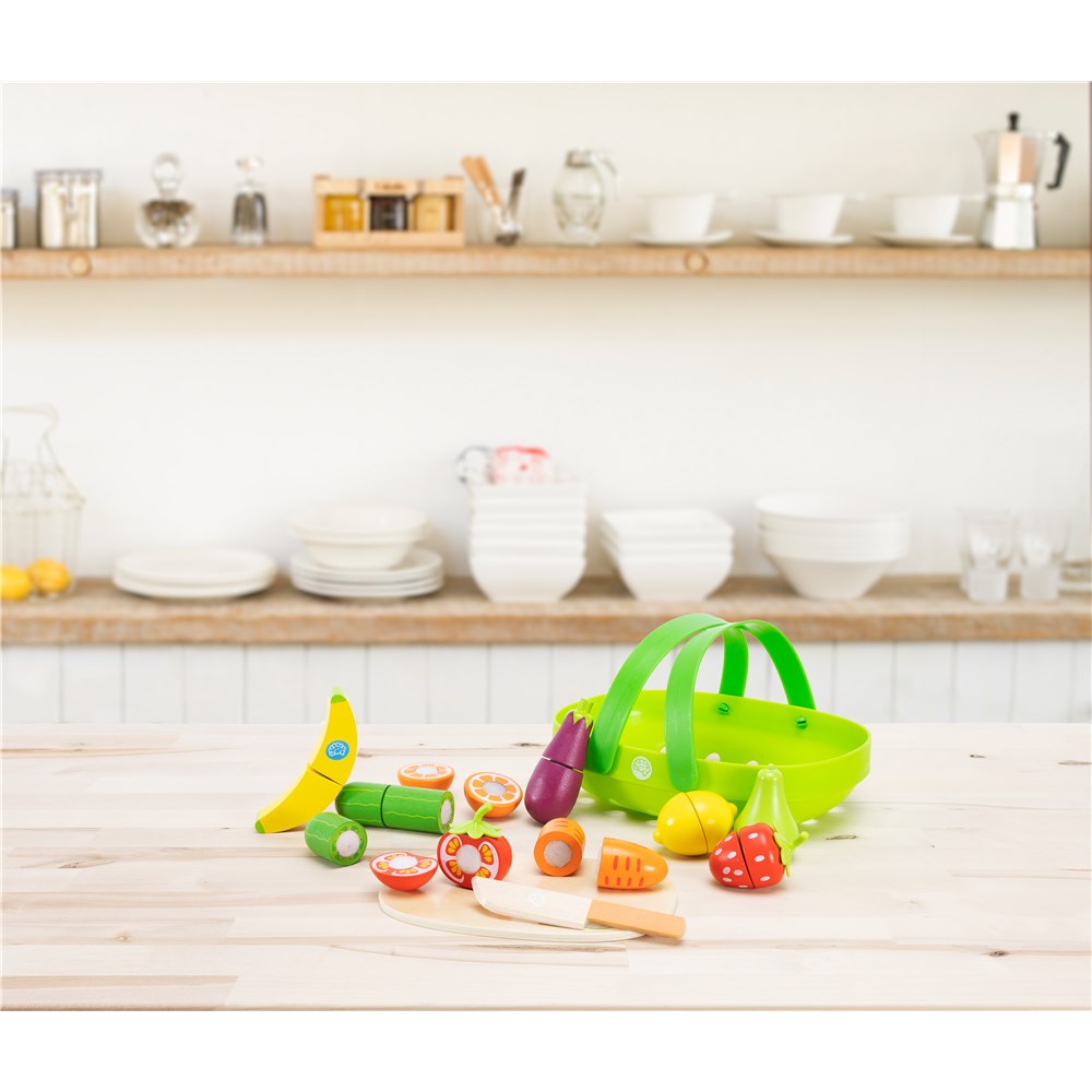 A colorful wooden toy kitchen scene with a green basket, banana, vegetables, and utensils arranged on a light wood table in front of a softly blurred kitchen background.