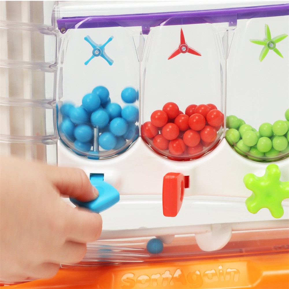 A close-up of a children's toy sorting machine with three clear bins filled with blue, red, and green balls, plus colored spinner pieces and a hand pressing a blue button to dispense a ball.