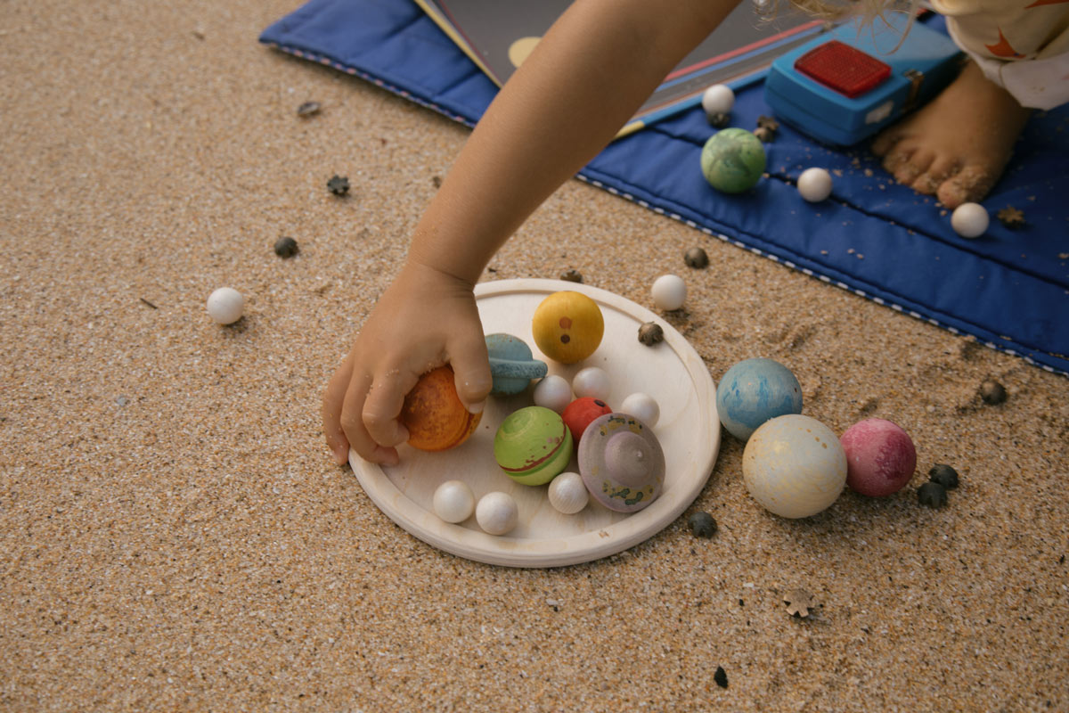 Child's hand holding a toy planet on a sand-like surface with more toy planets scattered around.