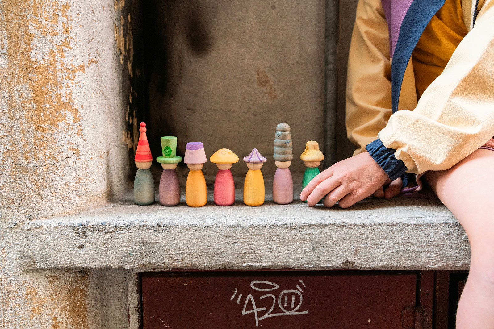 Colorful wooden figurines on a stone ledge with a child's hand reaching out.