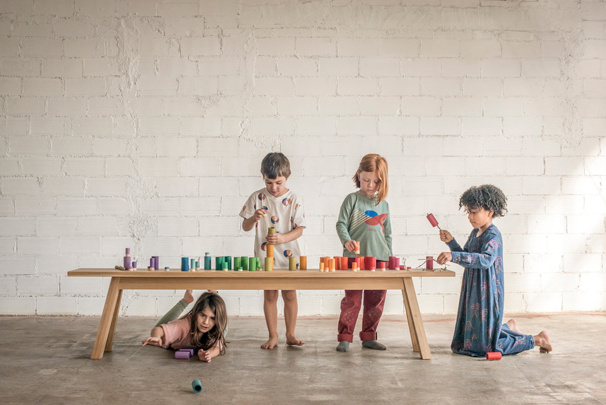 Four children playing with colorful toys around a wooden table against a white brick wall.