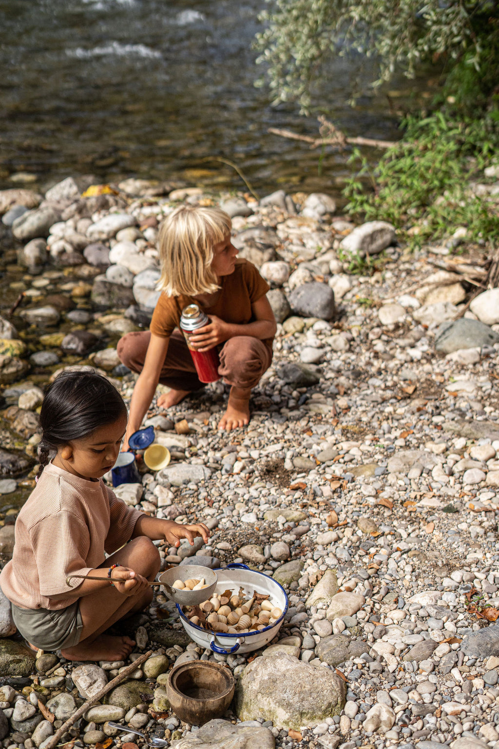 Two children by a river, one holding a red cup and the other with a bowl of stones.