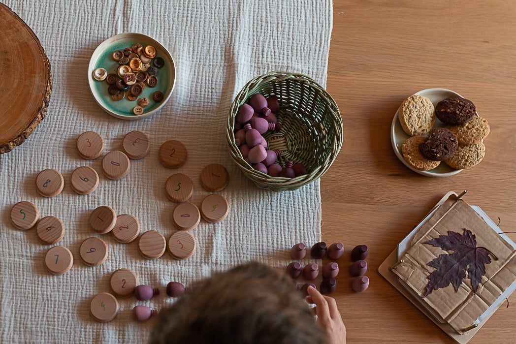Tabletop game with wooden pieces, a basket of purple stones, and a leaf-patterned box on a wooden surface.