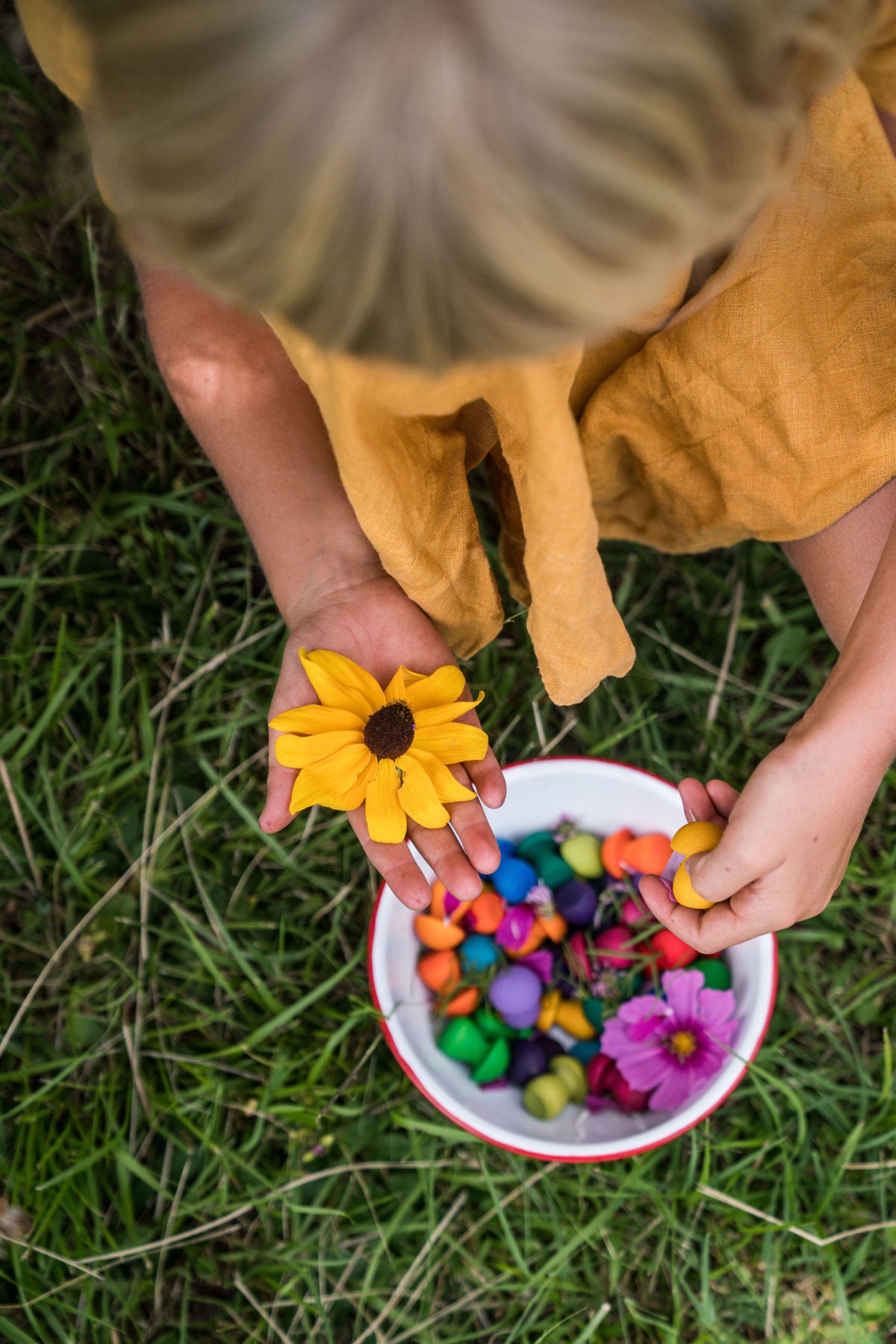 Child holding a bowl of colorful wooden toys and flowers on grass