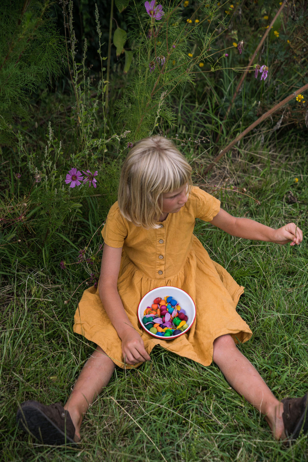 Young girl in a yellow dress sitting on grass with a bowl of colorful toys.