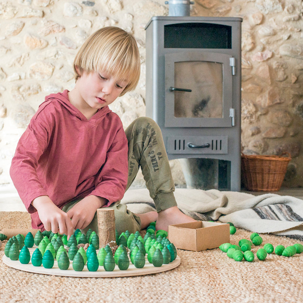 Child playing with green toys on a wooden tray in front of a stone wall and stove.