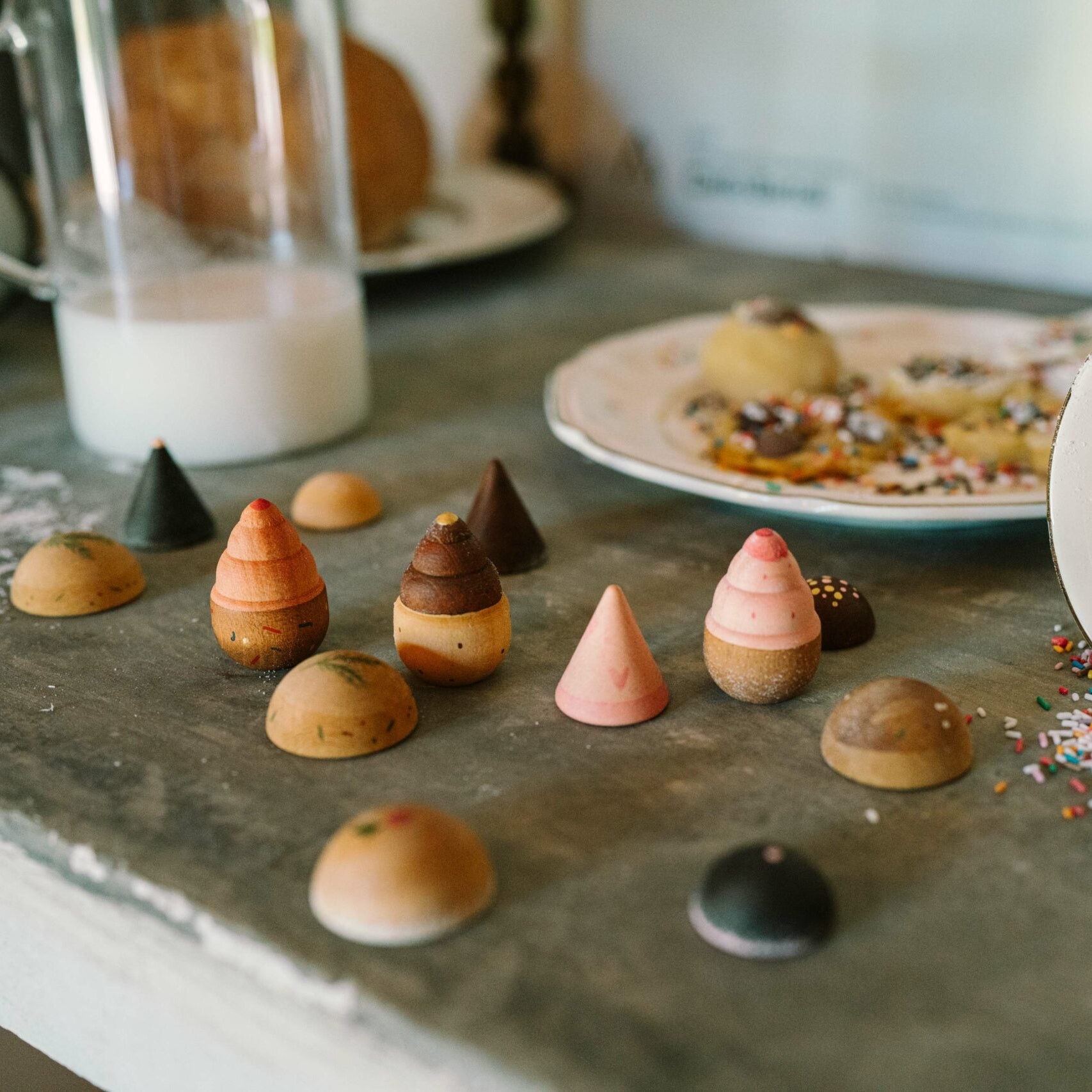 Colorful miniature pastries on a stone surface with a cup of milk and a bowl of sprinkles.