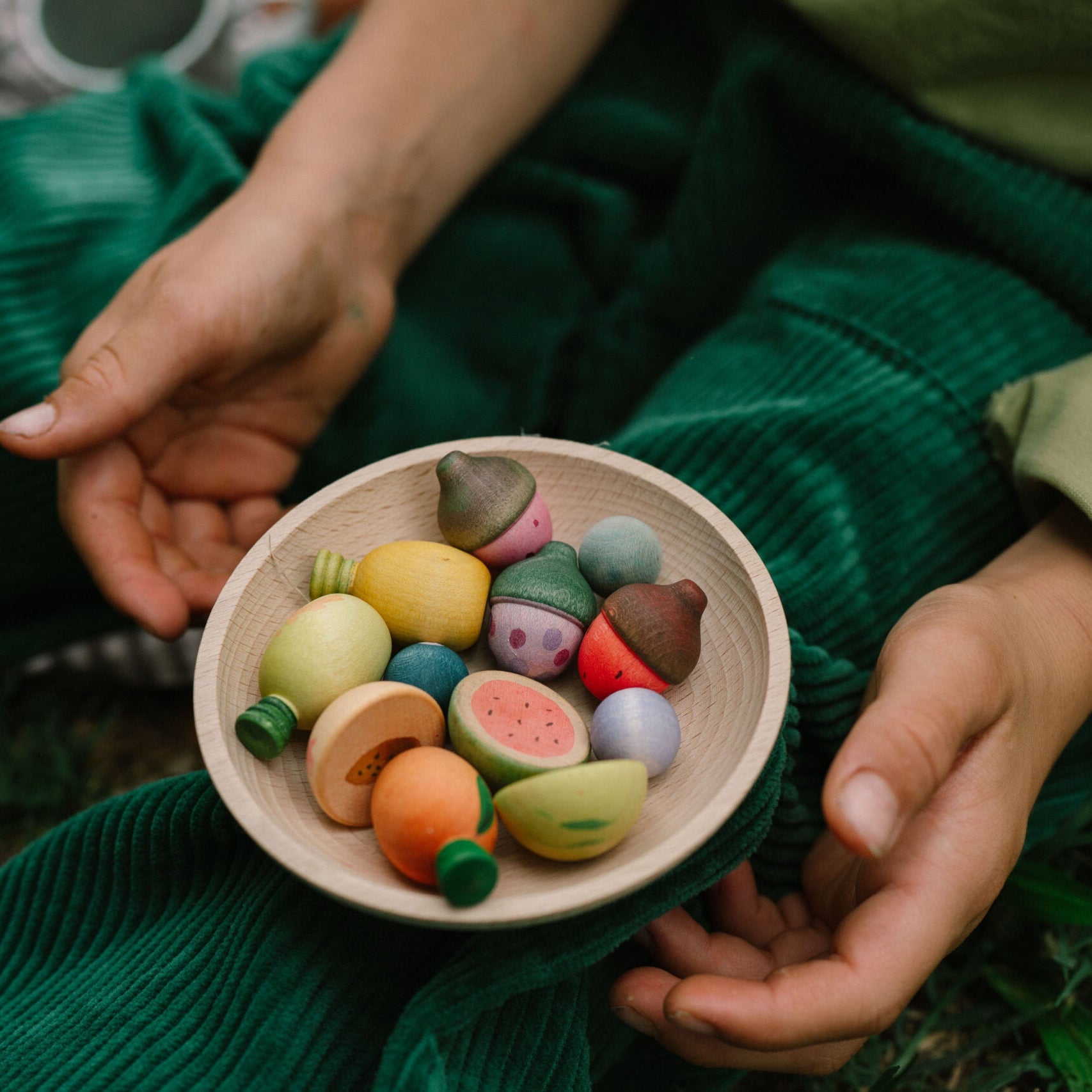 Person holding a bowl of colorful wooden figurines in a natural setting.