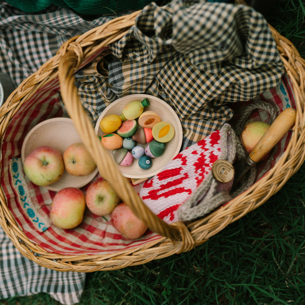 Wicker picnic basket with apples, candy, and a mug on a checkered blanket outdoors.