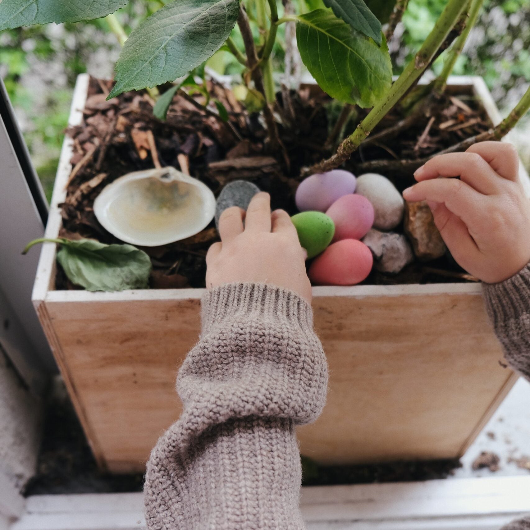 Children's hands interacting with colorful eggs in a planter box on a windowsill.