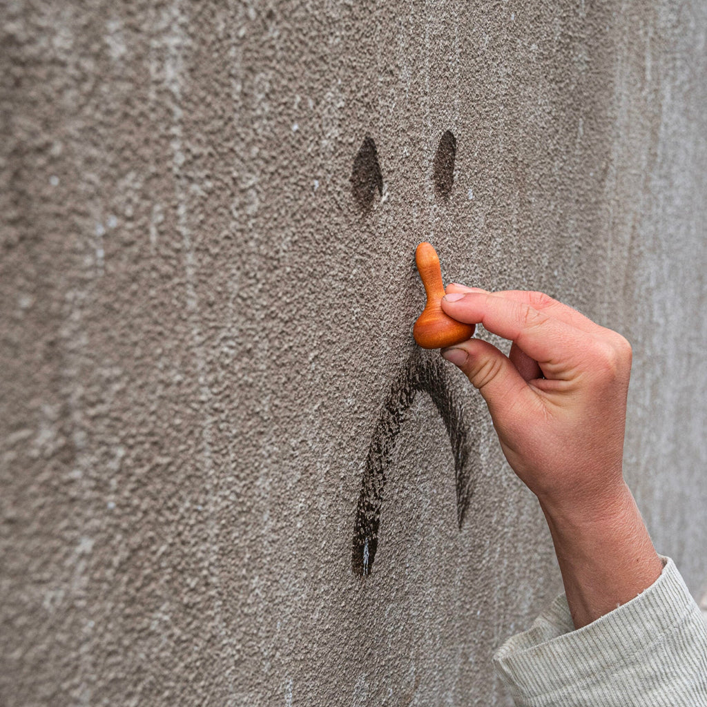 Hand holding a wooden spoon above a smiley face drawn on a textured wall.
